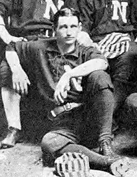 A black and white photograph of a man sitting on the floor wearing a dark baseball uniform with a white "N" on the chest. A striped cap rests at his feet.