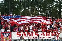 Soccer fans, dressed in red, cheer in bleachers as they hold a large American flag over themselves at a soccer match.