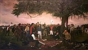 A group of men is gathered under a large tree.  One man lays on the ground under the trees, with his barefoot exposed.