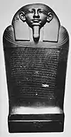 Black and white image of a dark stone coffin with a human face, the coffin stands upright facing the viewer.