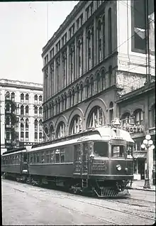 A train with two cars passing through a built-up downtown on tracks embedded in the street