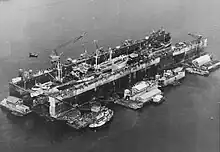 A floating drydock containing ships. It is surrounded by floating barges with workshops and a tugboat.