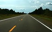 A straight roadway leading away to the horizon with trees on either side of the road