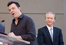 A man with black hair and slight stubble, wearing a black button-up shirt, speaking into a microphone. A man with gray hair, looking straight forward, and wearing suit, sits behind him.