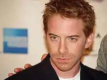 A young man with red hair, and stubble, looks slightly to his right. He is wearing a black jacket, and white shirt.