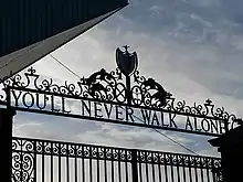 Design of the top of a set of gates, with the sky visible. The inscription on the gates reads "You'll Never Walk Alone".