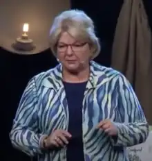 Short-haired blonde woman on a stage speaking into a headset microphone, wearing an animal stripe jacket with a bare lightbulb and draped cloth behind her