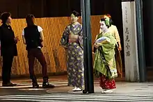 Two young women in kimono stood at a street corner at night. The taller woman wears a casual blue and yellow kimono; the smaller woman is dressed as an apprentice geisha, in a green kimono with green hair accessories and a red underkimono.