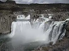 A wide, multi-stage waterfall, backed by dark cliffs, cascades into a rocky canyon