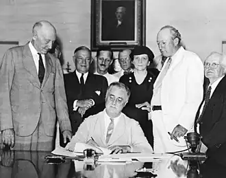 Franklin D. Roosevelt sits at a desk signing a document. Several people stand watching behind him.