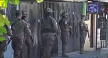 Soldiers in camouflage uniform, armed with rifles, and standing on a sidewalk in Soyapango