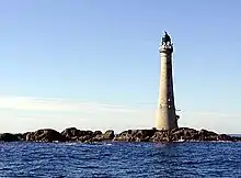 A tall lighthouse stands on low rocks under a blue sky.