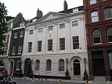 A four-storey, white bricked building, with two black doors. A flag pole is protruding from the centre of the building.