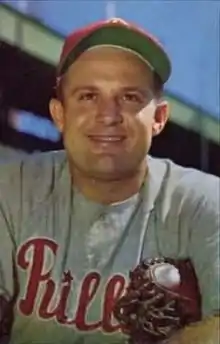 A man wearing a red baseball cap and a gray baseball jersey with "Phillies" written across the chest in red script smiles for the camera while clutching his baseball mitt under one arm.