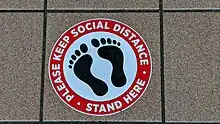 Circular sign with a silhouette of bare feet, with text in a red border that says "Please keep social distance • Stand here"