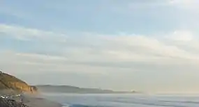 Finger of a mountain visible behind a cliff and on a beach with a partly cloudy sky