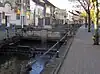 A river running between pavements with railings. Shops behind