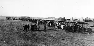 A slightly side-on front view of a line of early aircraft on a flat piece of land. People are standing with the aircraft, with a group standing in front of the machines. Several tent-like structures can be seen in the background.