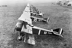 A side-on view of a line of several triplane aircraft. All are painted in dark colours, with white numbers on the side.