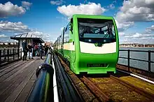 A narrow gauge train, in green and cream livery, runs along the pier railway in Southend on the right side of the photo. The sky is blue with white fluffy clouds. There are people walking along the pier on the left side of the photo.