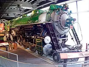 A green steam locomotive with a 4-6-2 wheel arrangement (four leading wheels, six driving wheels, and two trailing wheels) and its tender with a horse and cart in the foreground