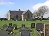 At the far end of a graveyard is a simple stone church, which appears L-shaped, and has a bellcote