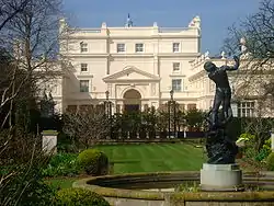 Symmetrical four-storey Neoclassical villa with an imposing pedimented entrance and balustrades around a valley roof surmounted by a small cupola and flanked by two-storey wings, the whole covered with stucco rendering painted pale pink. In front is a freshly mown lawn surrounded by plants and shrubs. In the foreground is a raised round stone pool with a bronze of a nude man being pulled into the water by a mermaid.