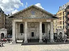 A sandy-coloured church with four large columns and a triangular pediment on top.