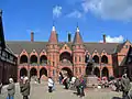 Stables, Eaton Hall, is directly north of the Chapel, in red brick, showing French late Gothic influence and the use of Tudor style half-timbering in the upper storey in the flanking ranges