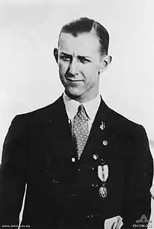 A close-up, mid-length black and white portrait of a young man in a dark suit. He is wearing a spotted tie, and has a medal on his left breast.