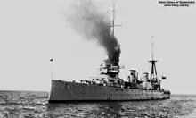 A large warship at anchor in calm waters. Black smoke is being emitted by the foremost exhaust funnel, and is obscuring the ship's forward masts