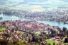 Aerial picture showing a small walled city dotted with red tile roofs and steeples; a wide river runs between it and another city. The farmlands on the far side are bathed in sunlight.