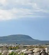 A grey-blue body of water separating smooth, large, light-coloured rocks in the foreground and a hazy green hill in the background, all under a blue sky with white clouds.