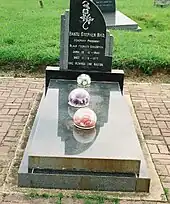 A grave and headstone. The structure is made from a shiny grey rock, and lacks ornamentation. Three flowers encased in circular glass baubles sit on top of it.