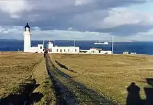 Stroma Lighthouse, tower on the left, support buildings to the right, viewed from a distance across grassland, up muddy tracks