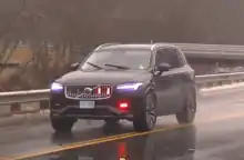 A new, black Volvo SUV driving in the rain, with red emergency lights activated while a firefighter responds to a call