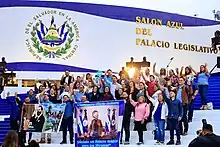 A crowd of people in front of the Legislative Assembly of El Salvador holding banners supporting Nayib Bukele and waving to a camera