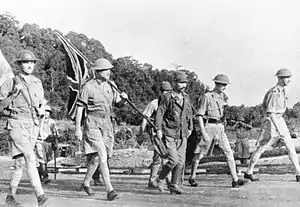 British soldiers holding flags march under escort of a Japanese soldier to surrender