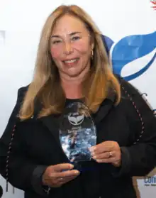 Picture of a middle-aged blonde woman. She's wearing a black sweatshirt and holds a crystal trophy with engraved letters to the camera. She stands in front of a white background with blue symbols and letters.