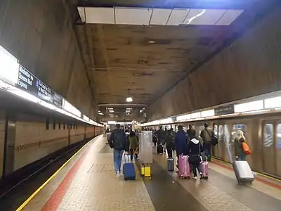 A view of passengers at the upper level, exiting an E train with luggage and heading for the AirTrain to JFK Airport