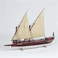 A side view of a model of a small galley with two masts rigged with lateen (triangular) sails. Its outrigger folded up and the oars stowed on the deck. The hull above the waterline is painted red with decorative details in gold and blue. The bow has a raised platform (rambade) armed with 3 small cannons.