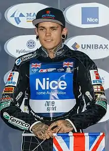 A man in his early twenties wearing a black baseball cap and speedway overalls with sponsors logos. He is sporting tattos on his necks and hands and his holding a miniature Union Jack in his right hand