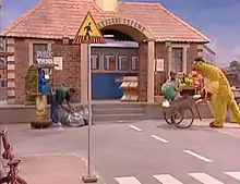 Television set, showing a large brick building on a city street; to the right, a large puppet is pushing a wheelbarrow and to the left, a man, next to a phone booth, is bending over several mail bags. Closer to the front of the image is a signpost, with a triangle that has a black figure and a yellow background, on top.