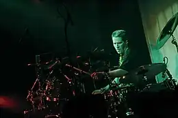 Drummer with crewcut, wearing black T-shirt, intently plays his large drum set in the spotlight.