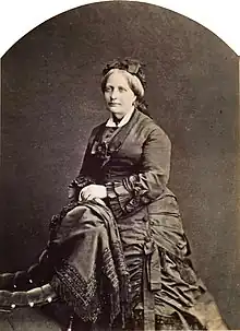 A photographic portrait of a woman with graying hair dressed in an elaborate dark mid-Victorian period dress and leaning against the back of an upholstered chair