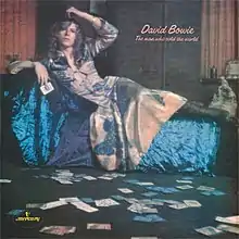 David Bowie in a blue dress laying on a couch with playing cards scattered across the floor