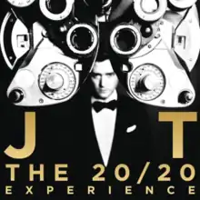 Black and white image on a black background. A blurred man is shown, wearing a suit and bowtie, looking at the camera from afar. In front of him is a phoropter and golden text reading "JT" and "THE 20/20 EXPERIENCE".