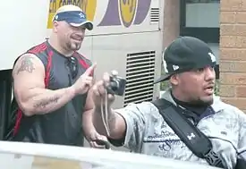 Two adult Latino males, one doing hand signs and another holding a camera.