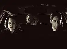 Two people are sitting in a car with a grotesque figure in the backseat. The scene is entirely in black and white footage.