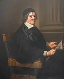 A man with a black cap and gown, seated.  He is holding an open book in his right hand and pointing to it with his left